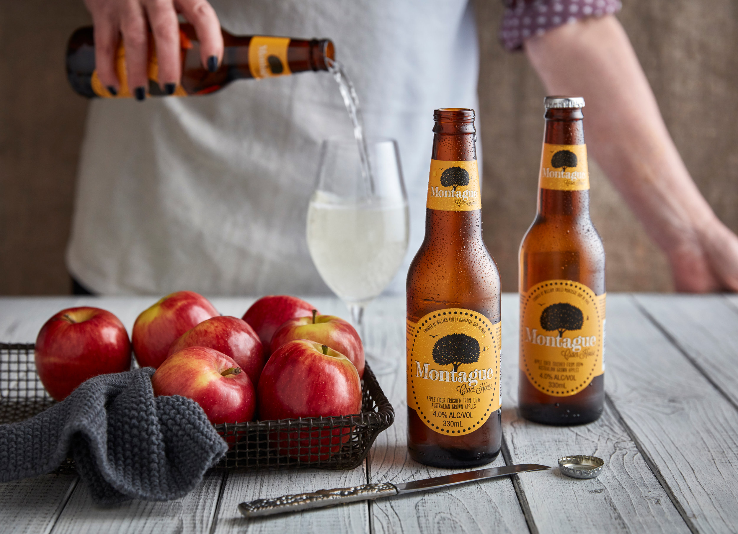 Pouring cider into a glass alongside apples and bottles of Montague cider on a white wooden table indoors, with a person visible.