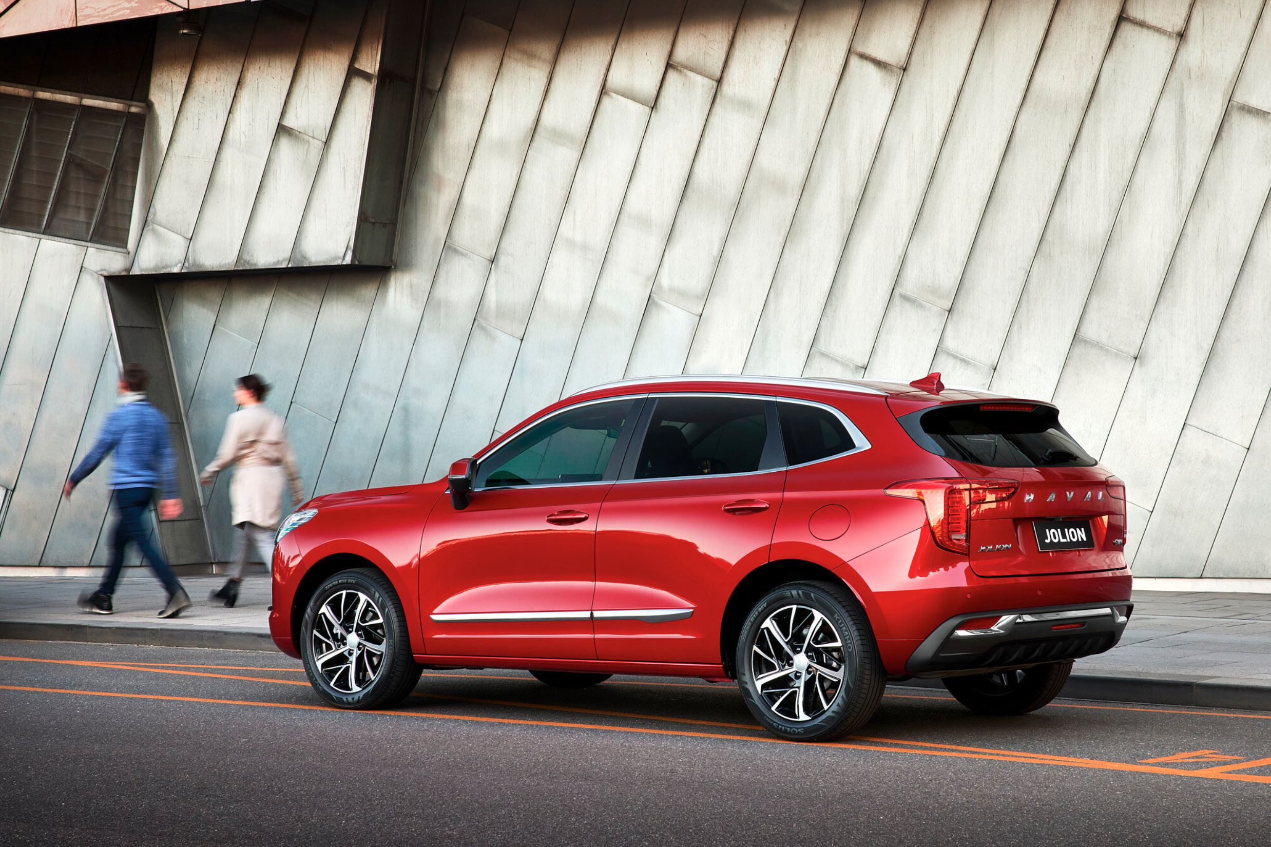 Red Haval Jolion compact SUV parked on the roadside in front of a modern building with two pedestrians walking in the background.
