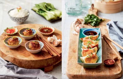 Overhead food spread showing pan-fried dumplings and meatballs on wooden boards with bowls of sauce and fresh greens.