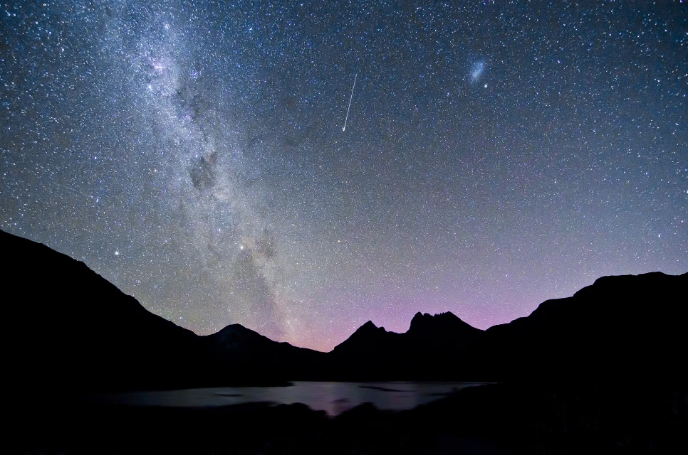 Night sky with Milky Way and shooting star over silhouetted mountains and lake creating a serene, celestial landscape.