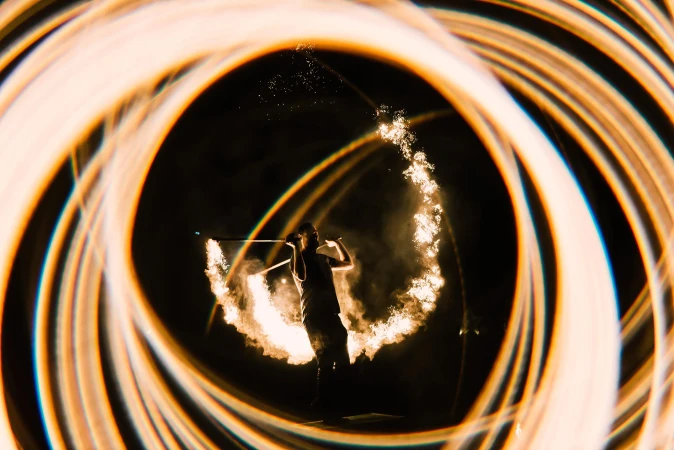 Silhouette of a person performing a fire dance at night, surrounded by swirling light trails in circular patterns around the performer.