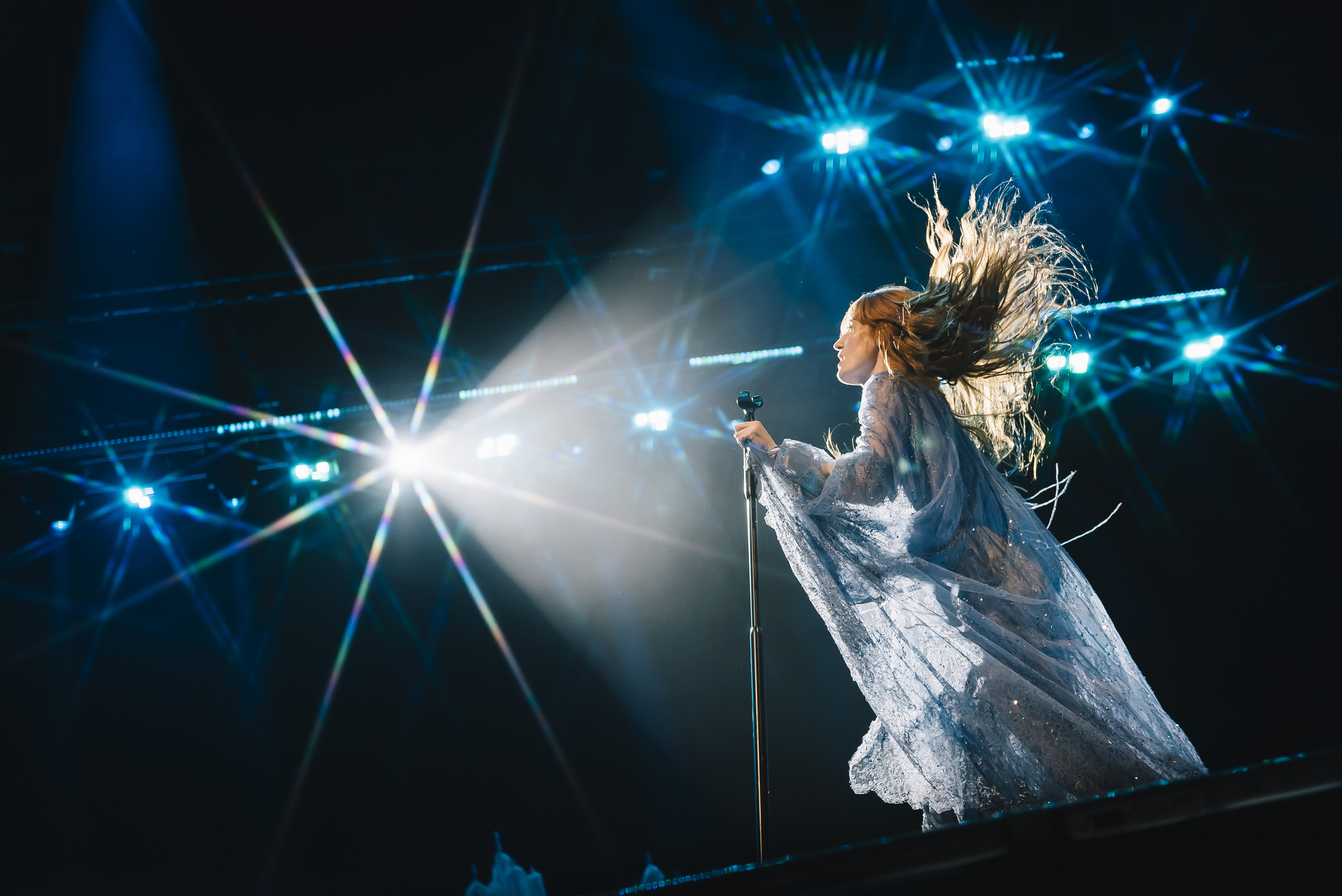 Singer performs on stage in shimmering gown with wind-blown hair, bathed in bright spotlights during a live music event.
