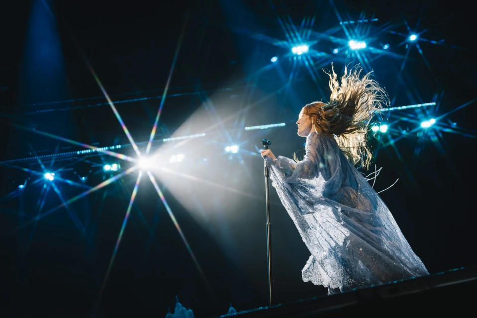 Singer performs on stage in shimmering gown with wind-blown hair, bathed in bright spotlights during a live music event.