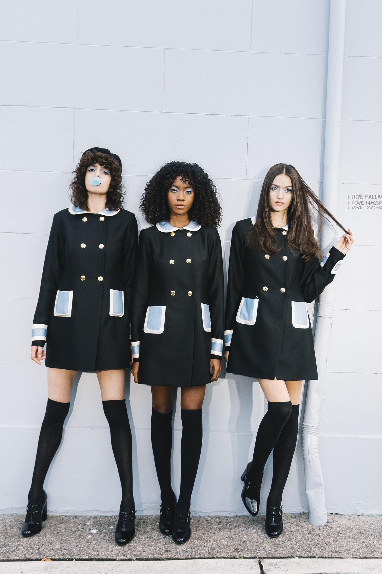 Three young women in mod-style black coats with white trim and knee socks pose against a pale textured wall with a vintage aesthetic.