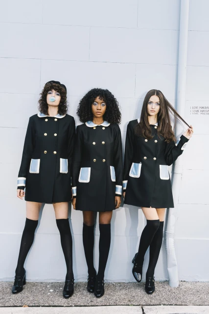 Three young women in mod-style black coats with white trim and knee socks pose against a pale textured wall with a vintage aesthetic.