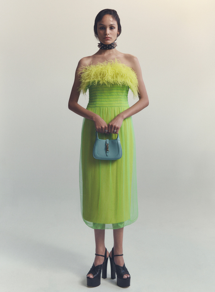 A model in a studio setting wears a lime green dress with a feathered neckline and chunky platform heels, holding a small, structured handbag.