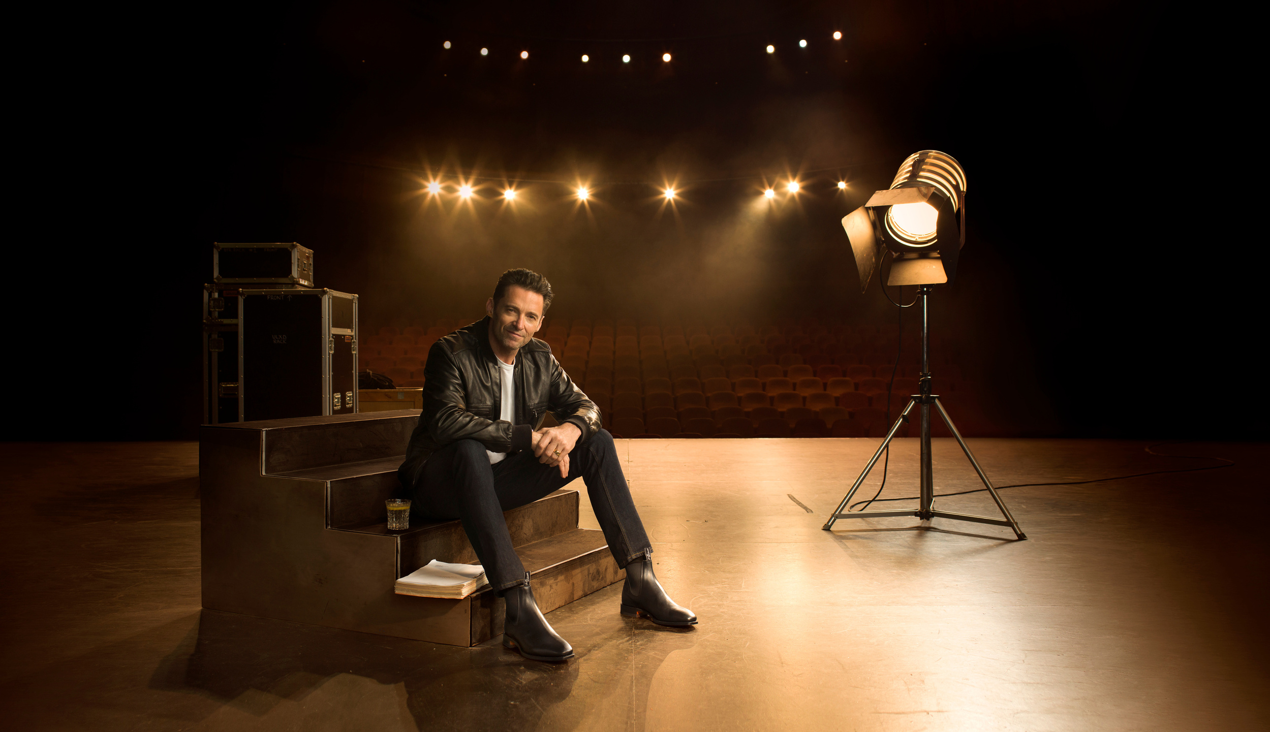 A casually posed man in a leather jacket sits on steps under stage lighting, adding depth and warmth to this celebrity and entertainment setting.
