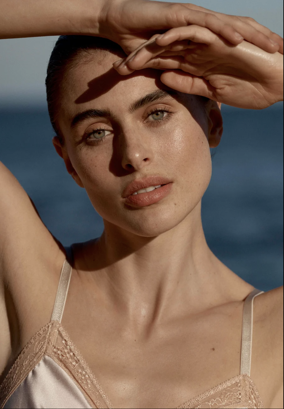 Close-up portrait of a woman with fair skin in a tank top against a blurry blue background, hand above head, in outdoor setting.