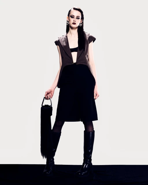 Full-length shot of a woman in a brown vest, black dress, and black knee-high boots, holding a black bag with fringe, against a white background.