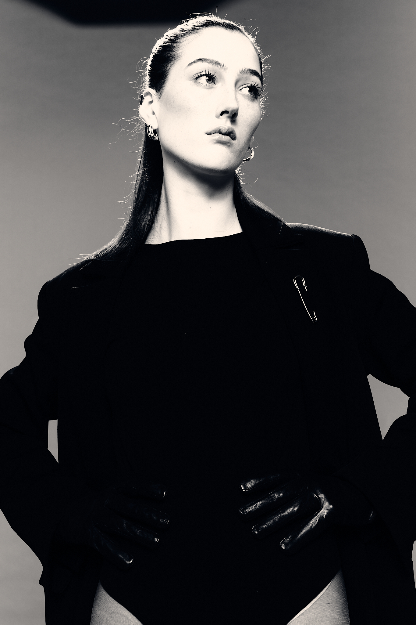Black and white fashion portrait of a woman with sleek hair wearing a black blazer and bodysuit, set against a neutral backdrop.