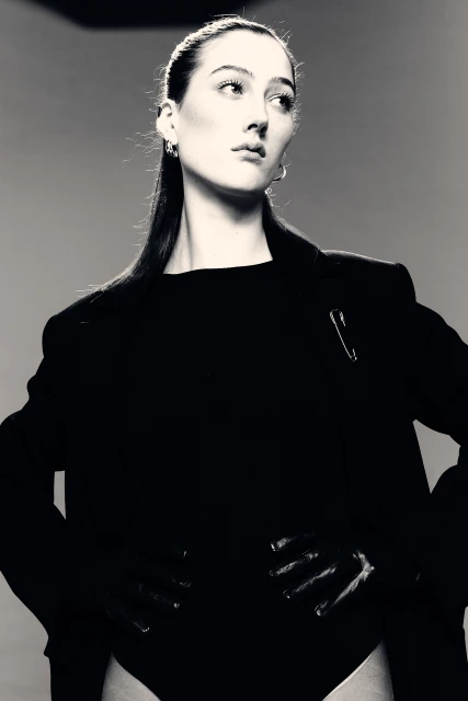 Black and white fashion portrait of a woman with sleek hair wearing a black blazer and bodysuit, set against a neutral backdrop.