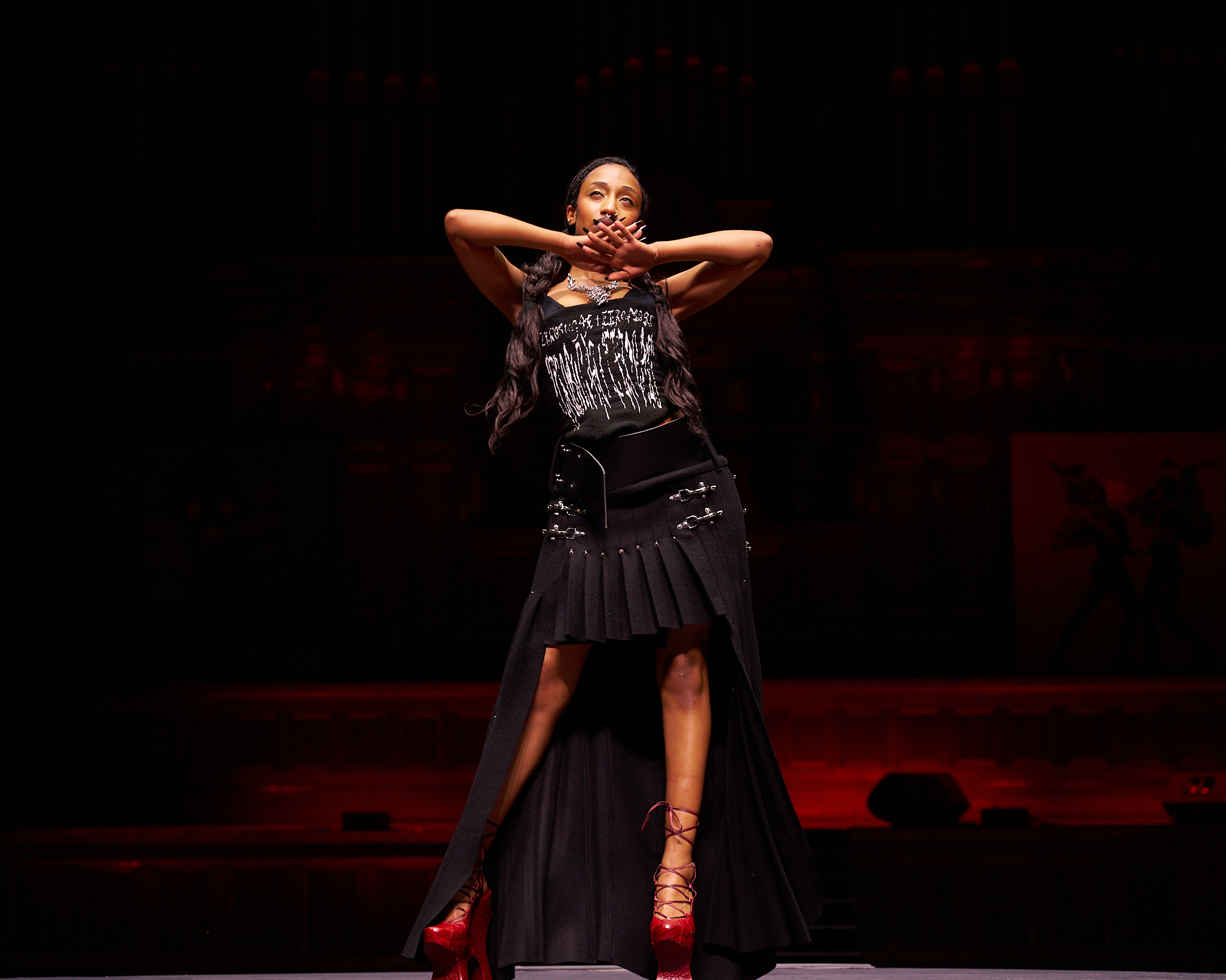 A woman in a black outfit and red platform shoes poses on a stage during a runway show captured by Zach Dean.