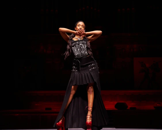 A woman in a black outfit and red platform shoes poses on a stage during a runway show captured by Zach Dean.