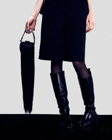 Model in black shorts and knee-high leather boots holding a unique long black fringe handbag against a white background
