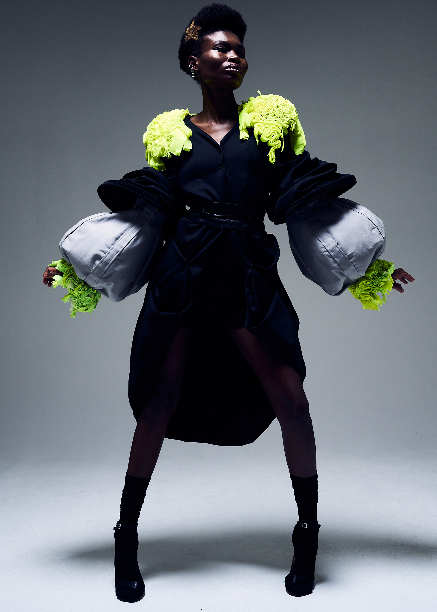 Full-length shot of a Black model wearing a black dress with oversized silver sleeves and chartreuse trim, posing in a studio setting.
