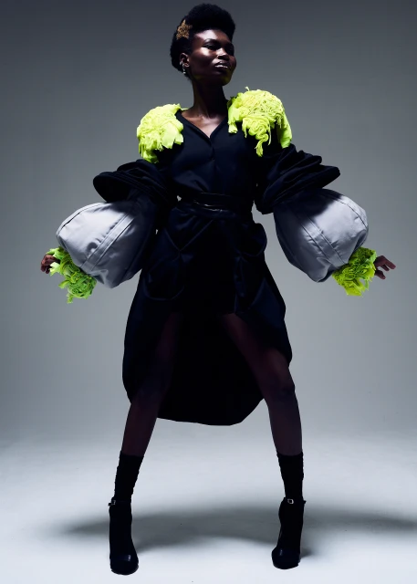 Full-length shot of a Black model wearing a black dress with oversized silver sleeves and chartreuse trim, posing in a studio setting.