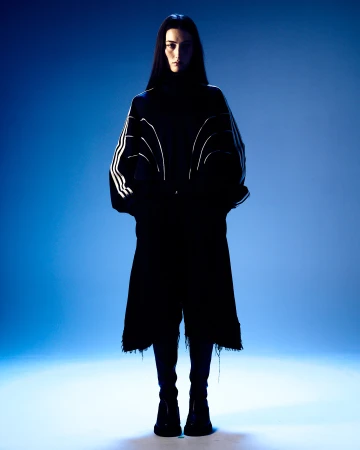 Model with long dark hair wearing a dark coat with white striped sleeves against a blue studio background