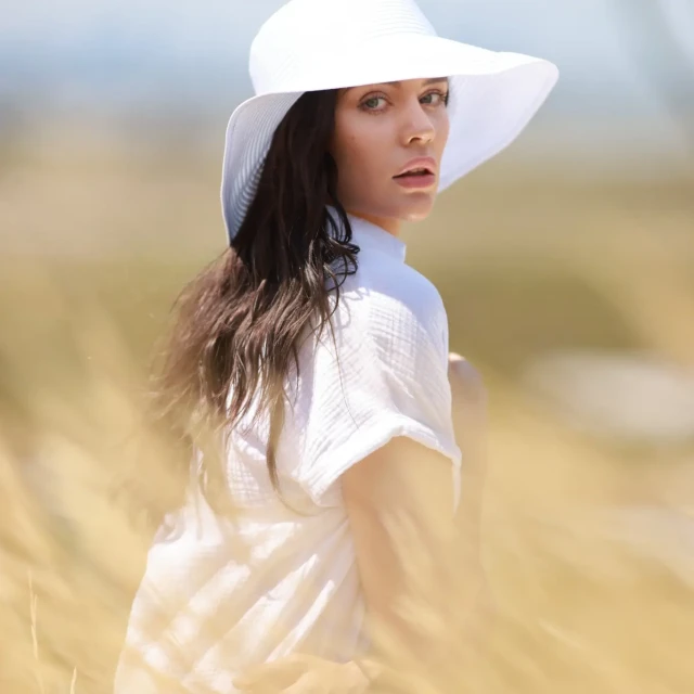 Brunette woman wearing a white hat and shirt looks over her shoulder in a sunlit field of tall golden grass in an outdoor setting.