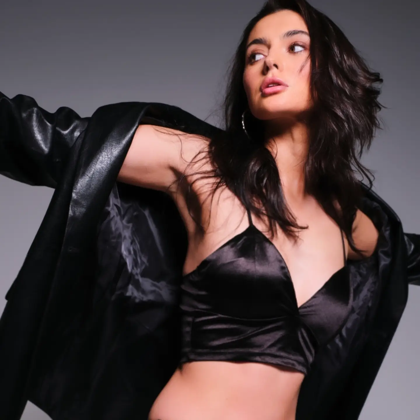 Brunette woman poses in a black satin top and open leather jacket against a grey backdrop in a studio portrait.