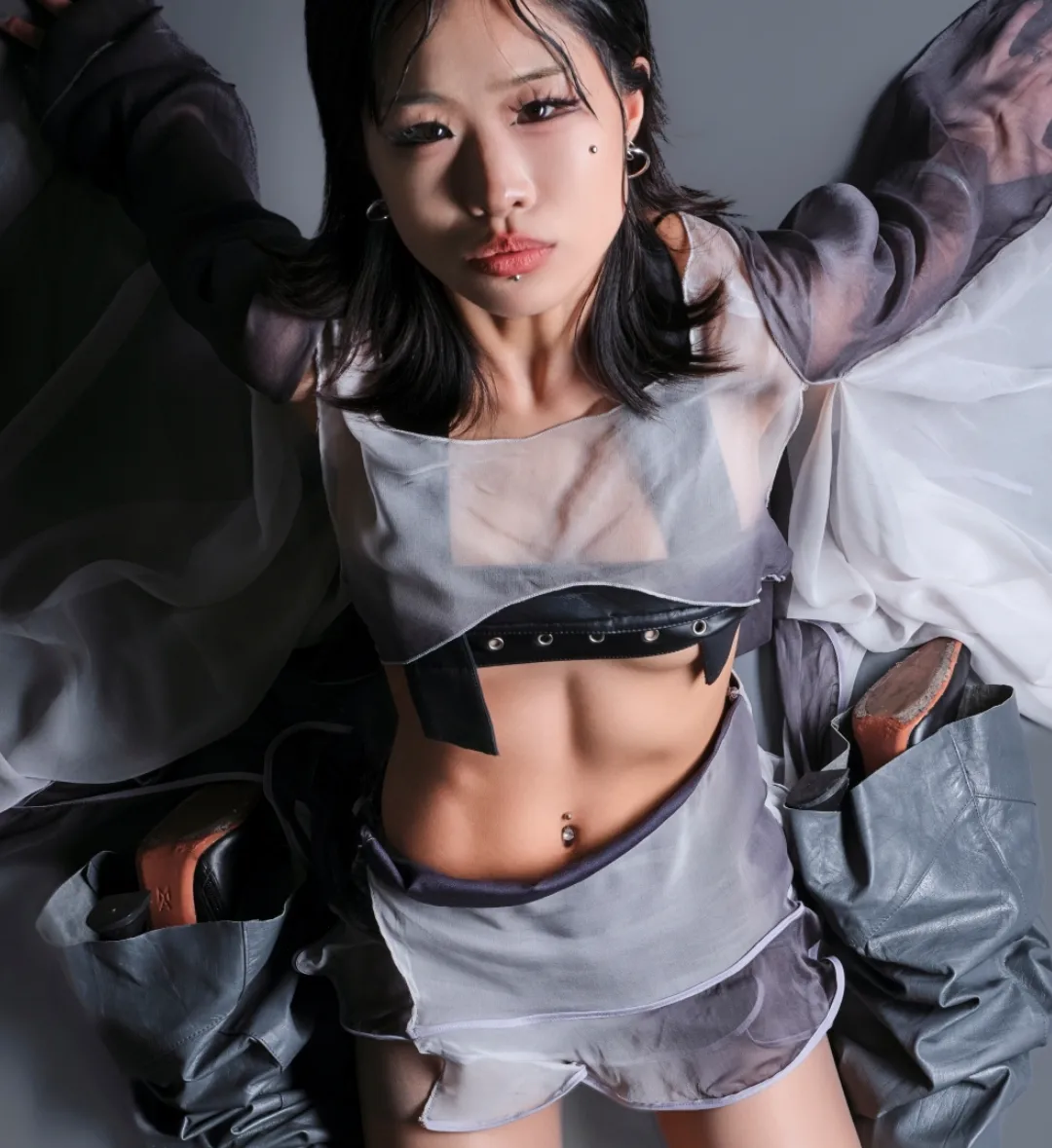 Young woman in sheer grey top and shorts lies on grey background, posing with shoes at her sides, looking at the camera.