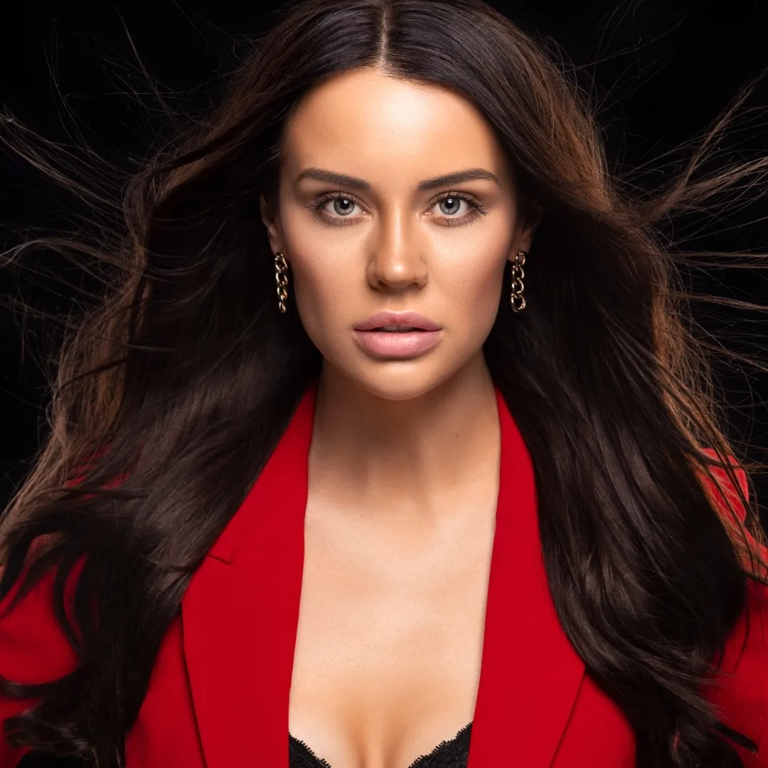 Portrait of a woman with long brown hair wearing a red blazer and gold earrings against a dark background in a studio setting.