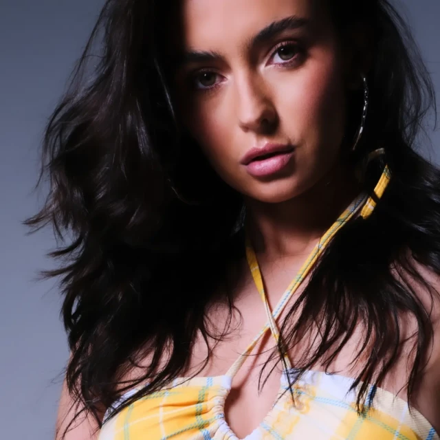 Portrait of a brunette woman with wavy hair wearing a yellow plaid halter top and hoop earrings in a studio setting with soft lighting.