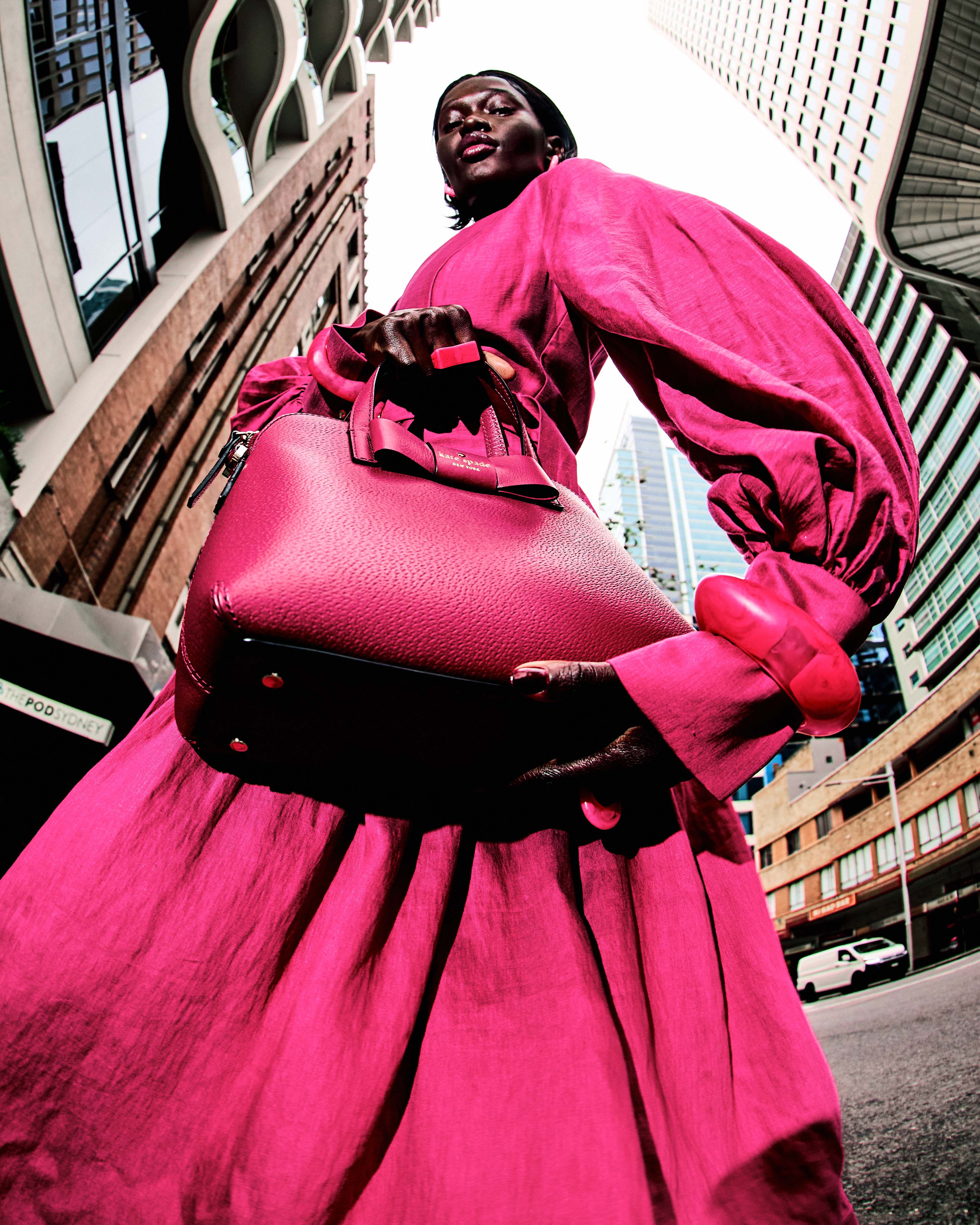 Fashion photography of an adult Black female model wearing a pink dress and holding a purse in an urban cityscape with tall buildings.