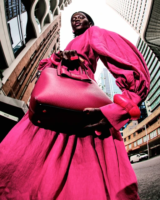 Fashion photography of an adult Black female model wearing a pink dress and holding a purse in an urban cityscape with tall buildings.