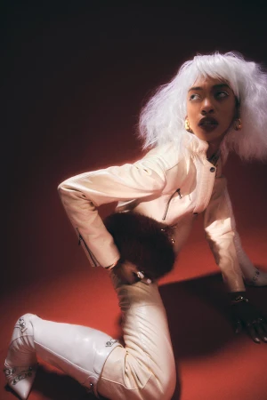 Model with white bob wig wearing a cream leather jacket and matching pants kneeling against a warm reddish background
