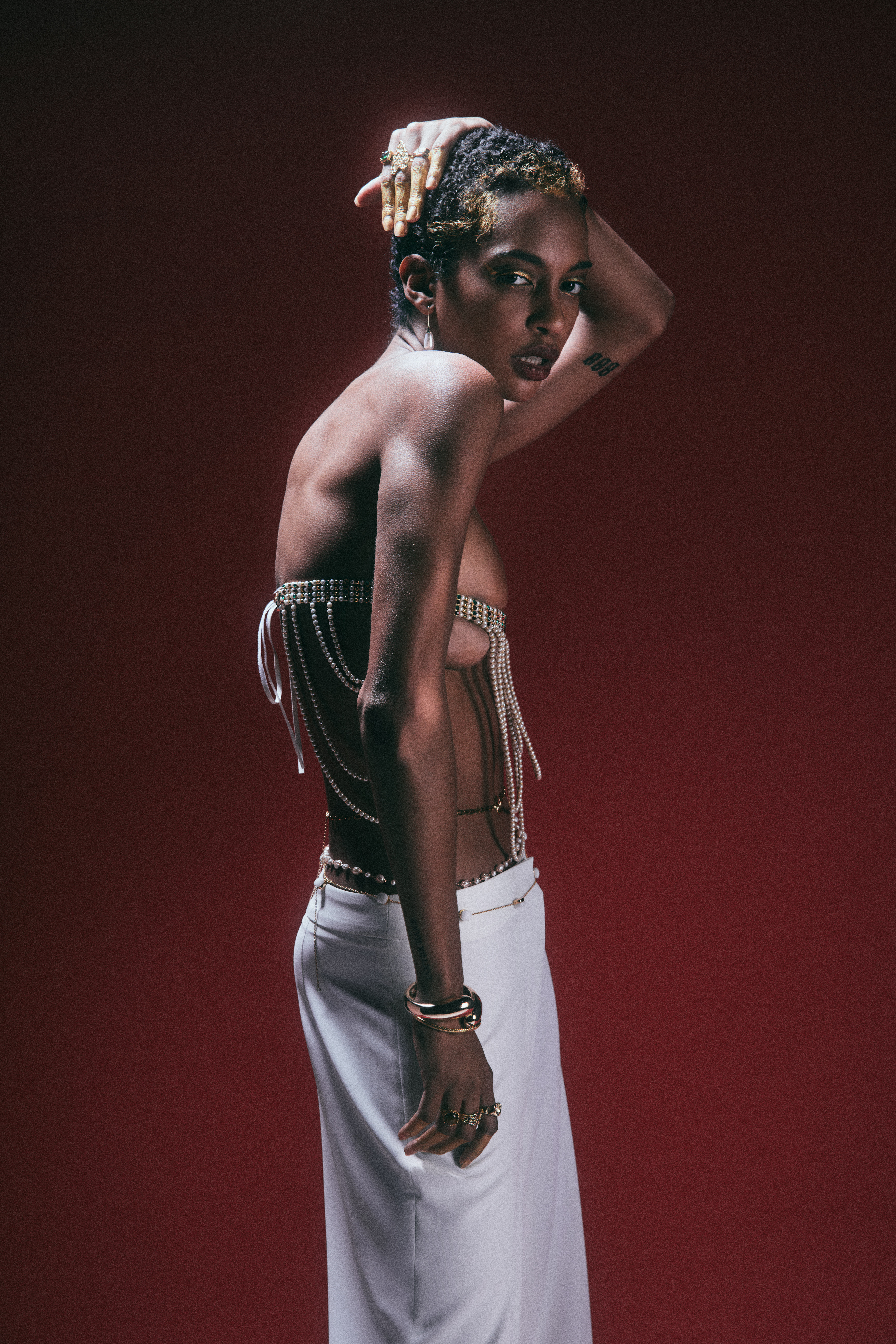 Woman with short textured hair poses in a chain top and white skirt against a red background with soft lighting and a serious expression.