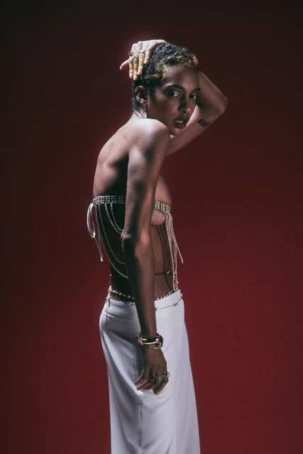 Woman with short textured hair poses in a chain top and white skirt against a red background with soft lighting and a serious expression.