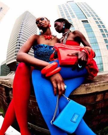 Two dark-skinned women in bright fashion outfits, posing with handbags and jewelry, sitting on a brick wall against a skyscraper backdrop.