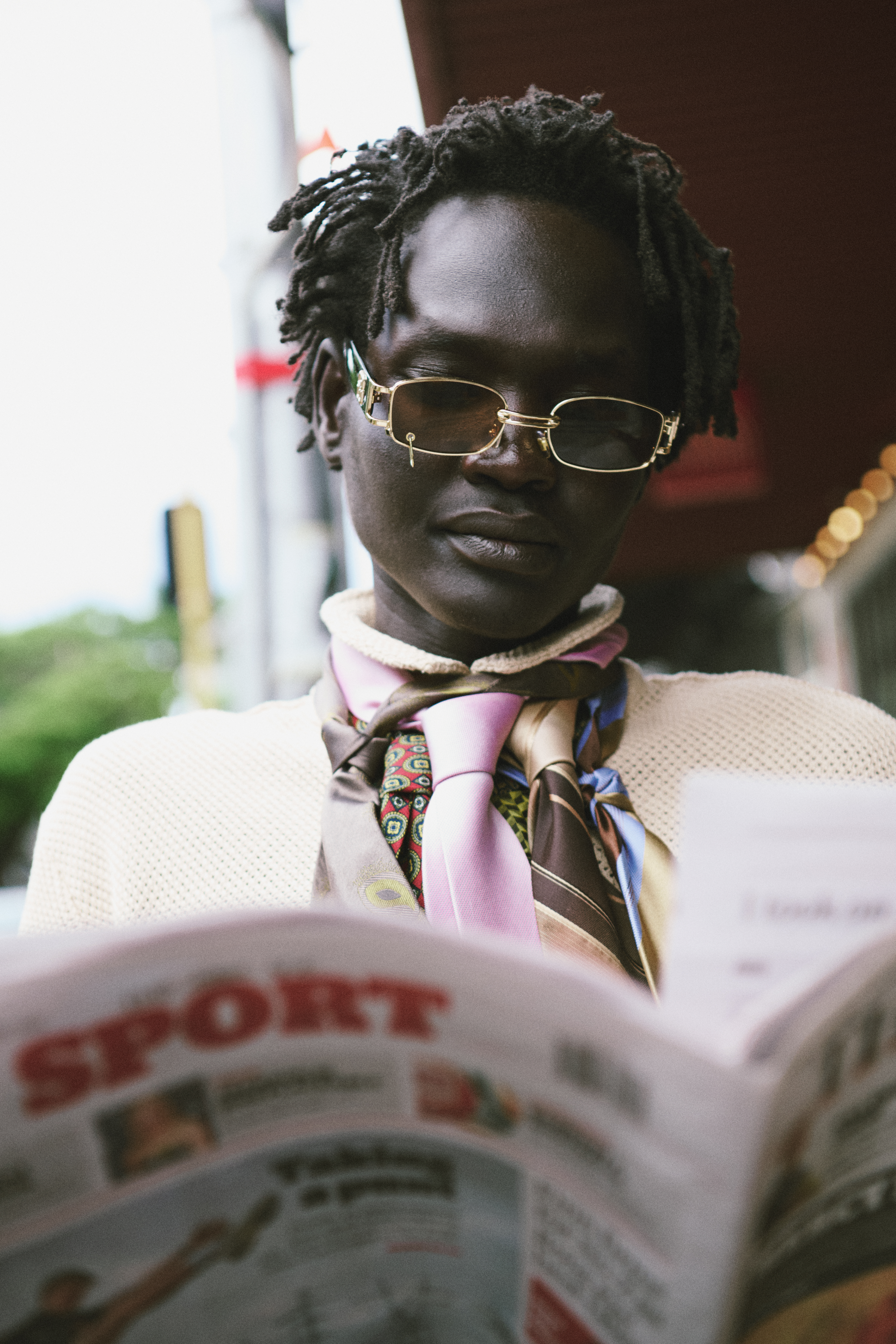 Person with dark skin and dreadlocks, wearing glasses and scarf, reading a newspaper in an outdoor urban setting.