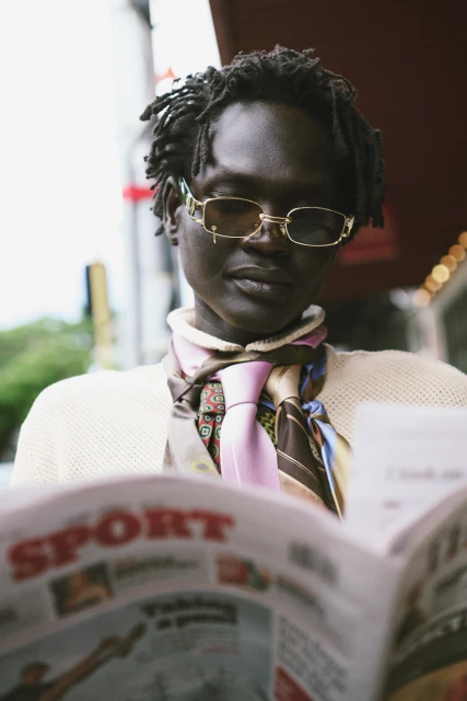 Person with dark skin and dreadlocks, wearing glasses and scarf, reading a newspaper in an outdoor urban setting. 