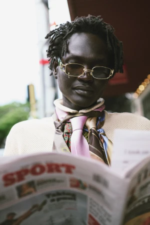 Person with dark skin and dreadlocks, wearing glasses and scarf, reading a newspaper in an outdoor urban setting. 