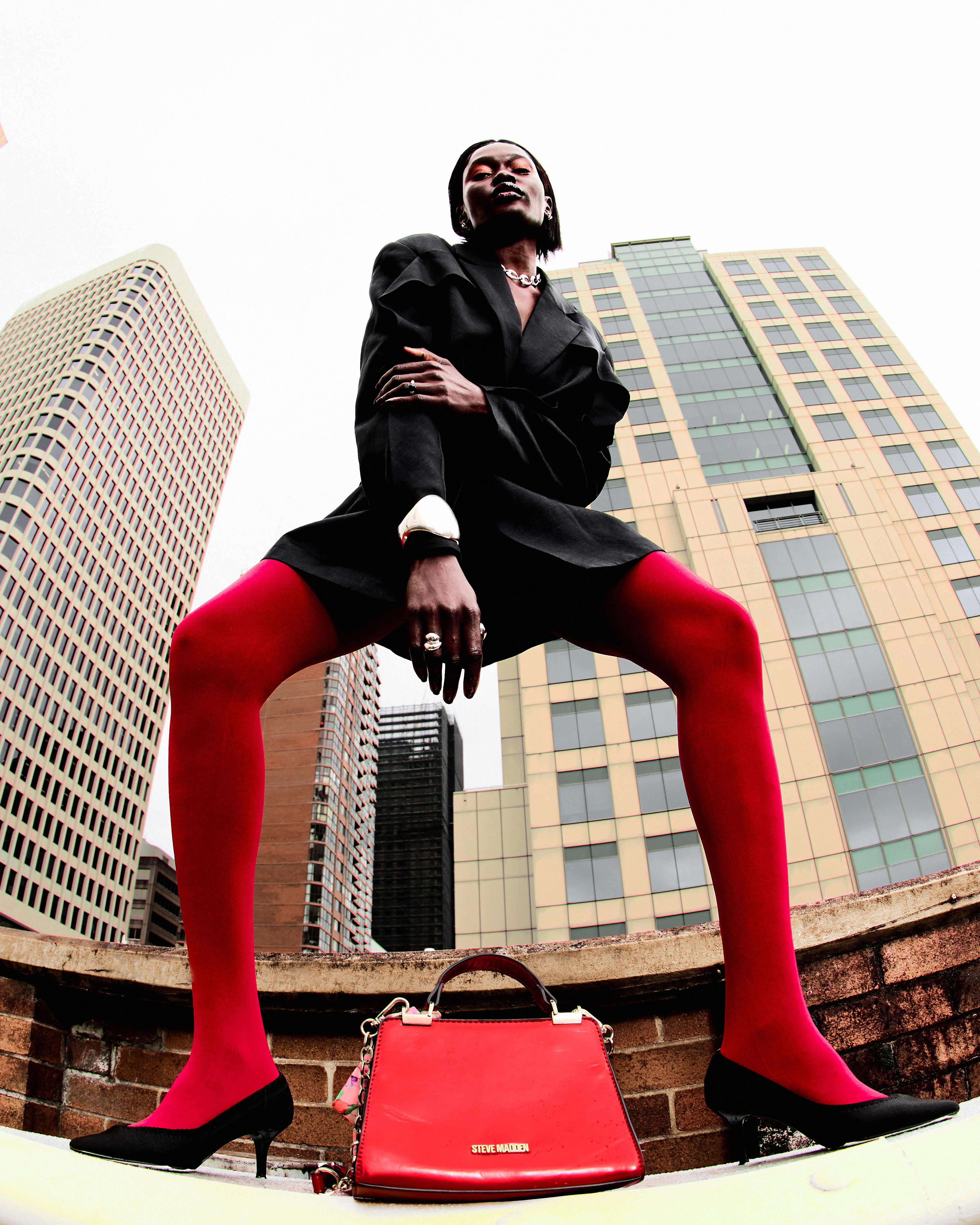 Model wearing a black blazer and red tights standing wide over a red handbag with tall buildings in the background