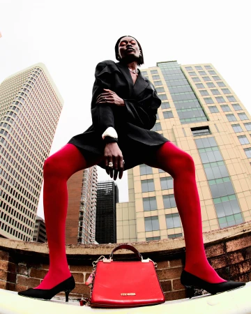 Model wearing a black blazer and red tights standing wide over a red handbag with tall buildings in the background