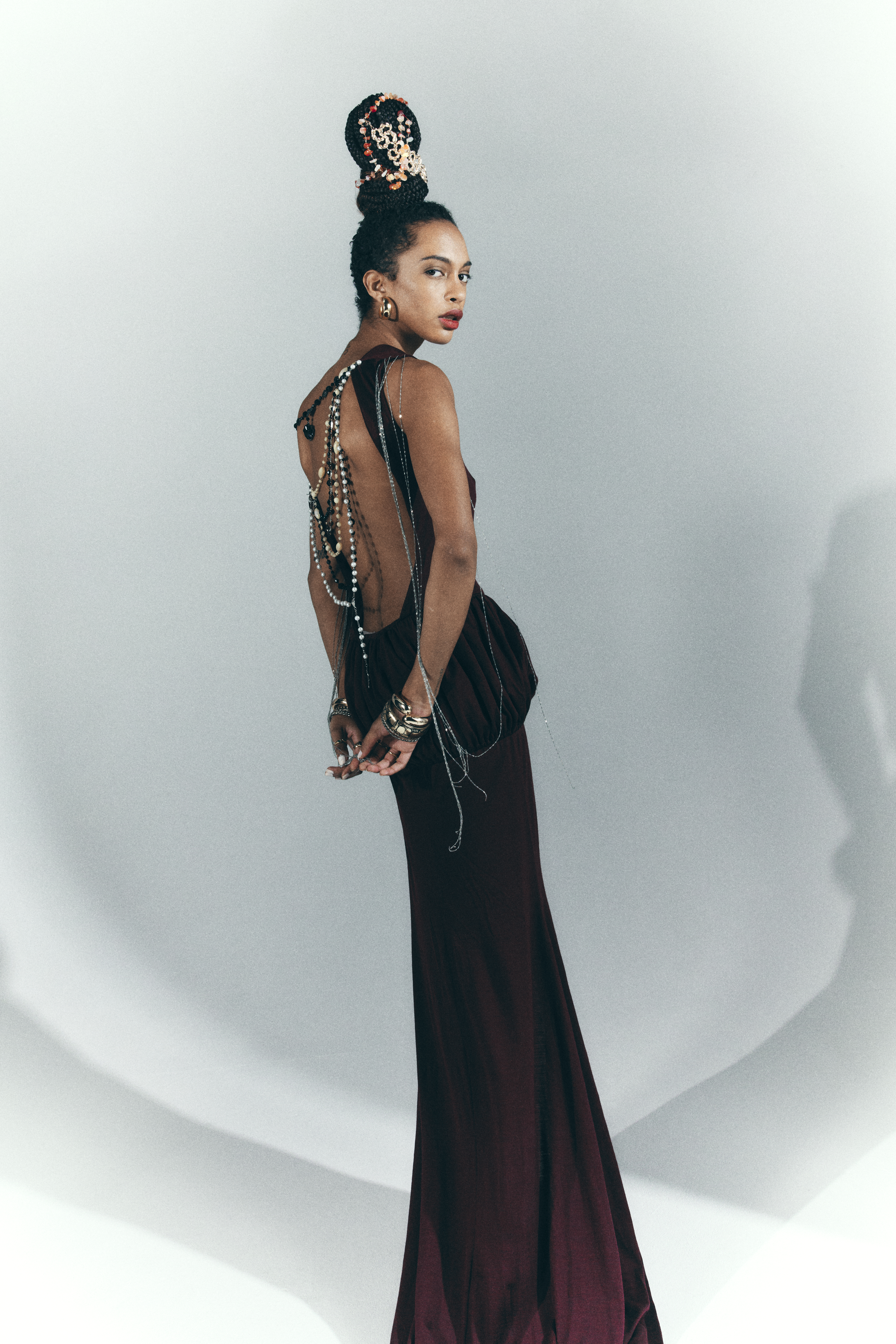 Fashion shot of a woman in a burgundy dress and jewelry, looking over her shoulder against a white backdrop in a photo studio.