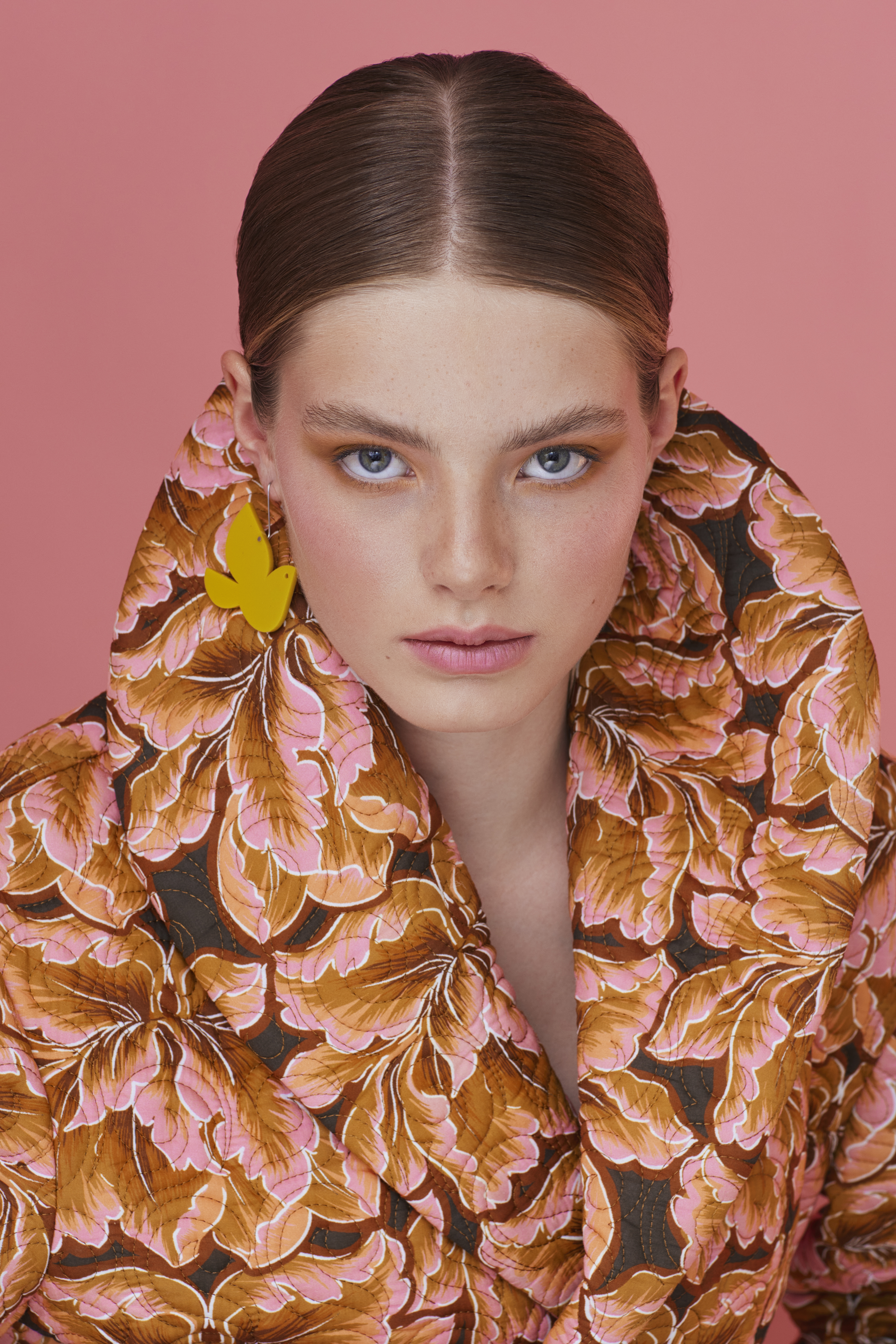 Close-up portrait of a young adult woman with light skin, wearing a leaf-patterned jacket and yellow earrings against a pink backdrop.