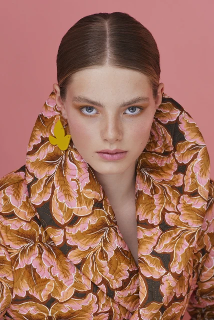 Close-up portrait of a young adult woman with light skin, wearing a leaf-patterned jacket and yellow earrings against a pink backdrop.