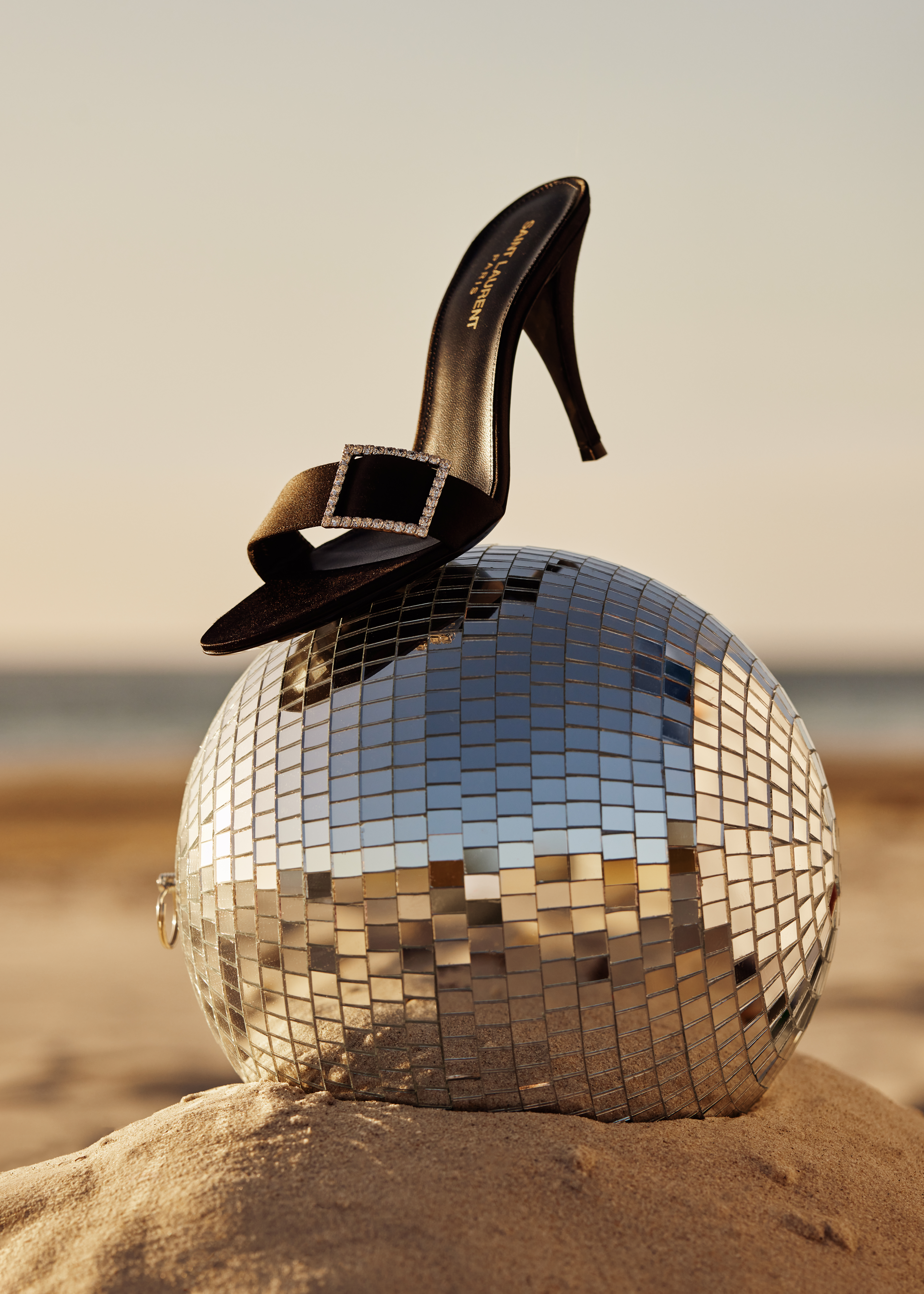 Black high-heel mule by Saint Laurent resting atop a mirrored disco ball on sand at the beach against the sky at golden hour.