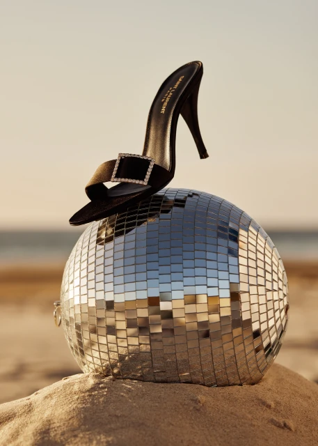 Black high-heel mule by Saint Laurent resting atop a mirrored disco ball on sand at the beach against the sky at golden hour.