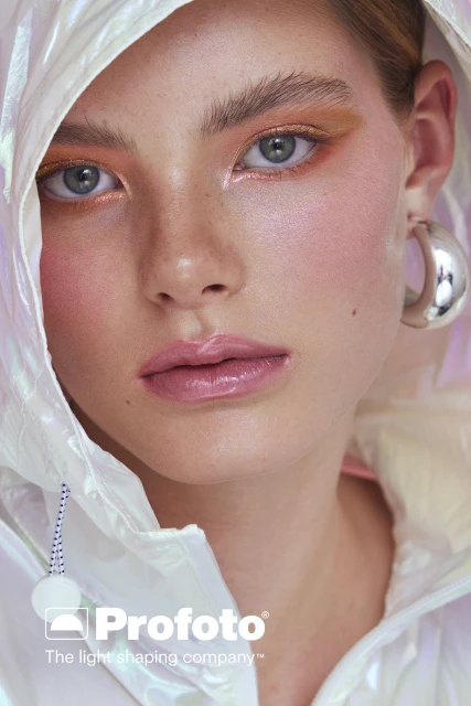 Close-up portrait of a young adult woman with blue eyes and colorful makeup wearing a white iridescent hooded jacket in a studio setting.