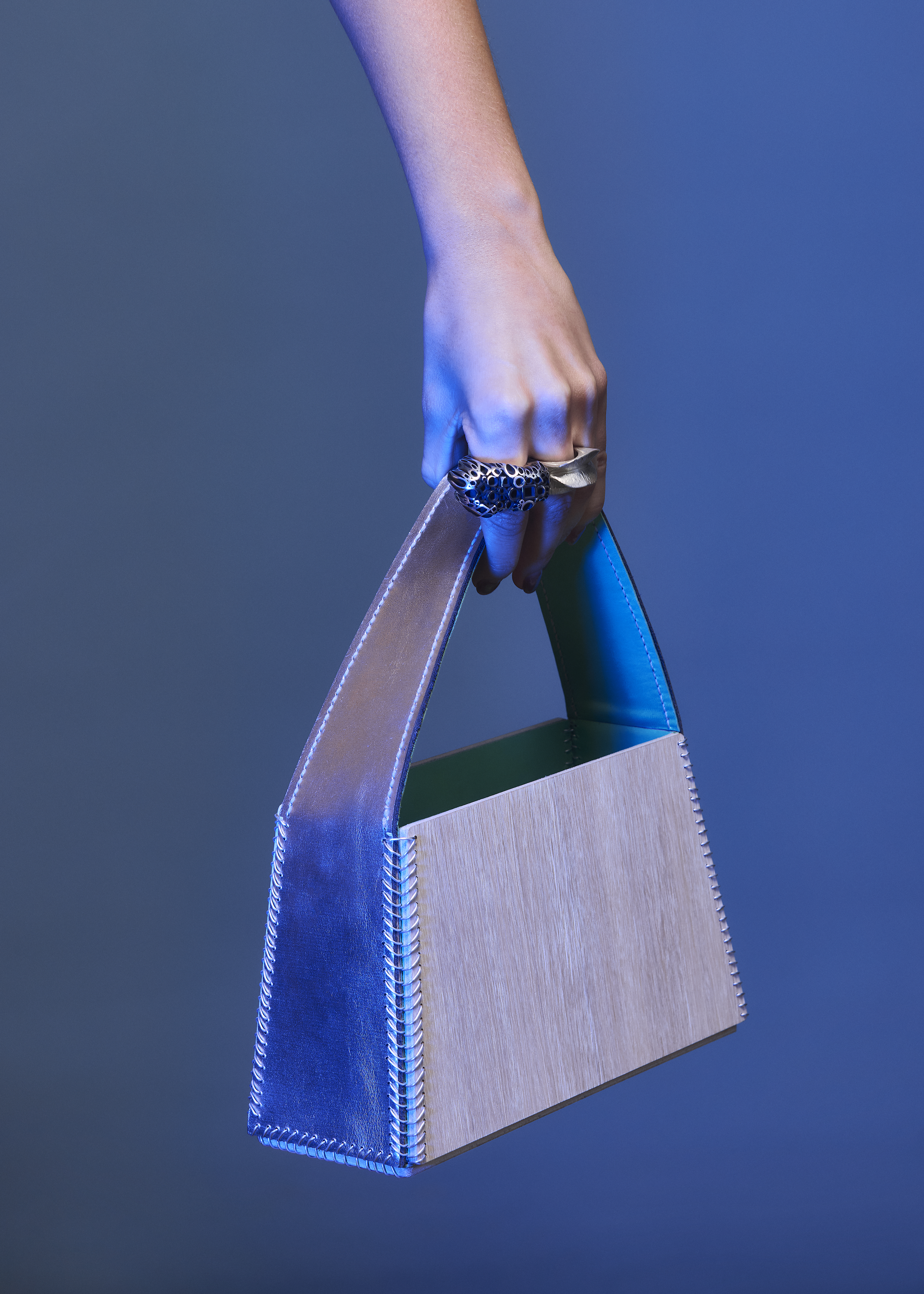 Close-up of an adult's hand holding a modern handbag with metal rings and a structured, geometric design against a blue background.
