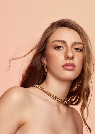 Close-up portrait of a woman with freckles wearing a thick gold chain necklace and ornate earrings against a solid peach background.