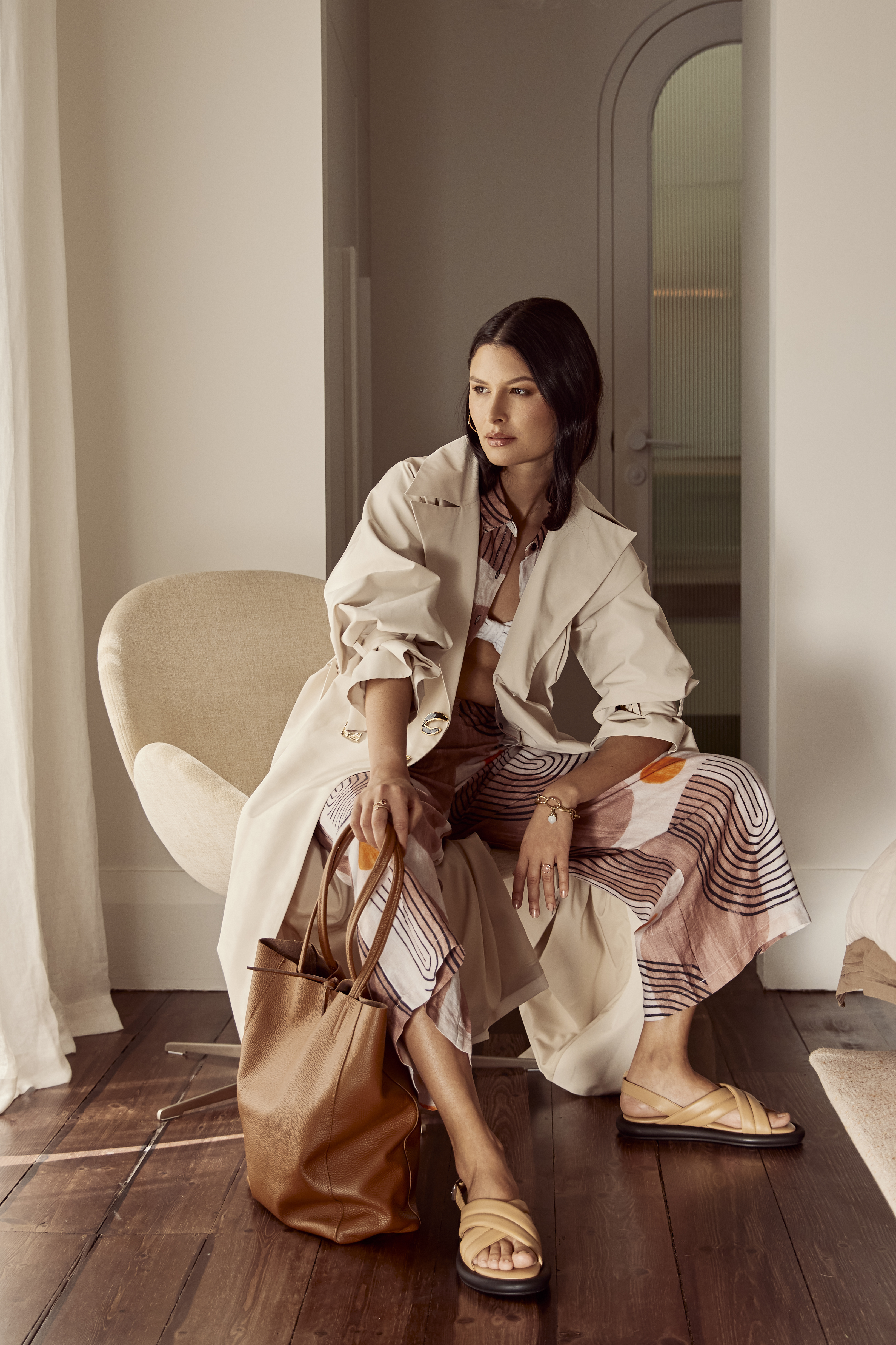 A woman is sitting in a chair wearing a patterned outfit, tan trench coat, and sandals while holding a brown leather tote bag indoors.