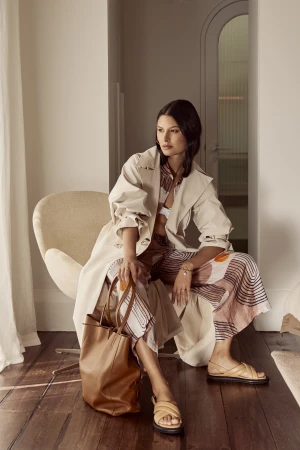 Woman in patterned trousers and cream trench coat seated in a white chair holding a brown leather tote bag