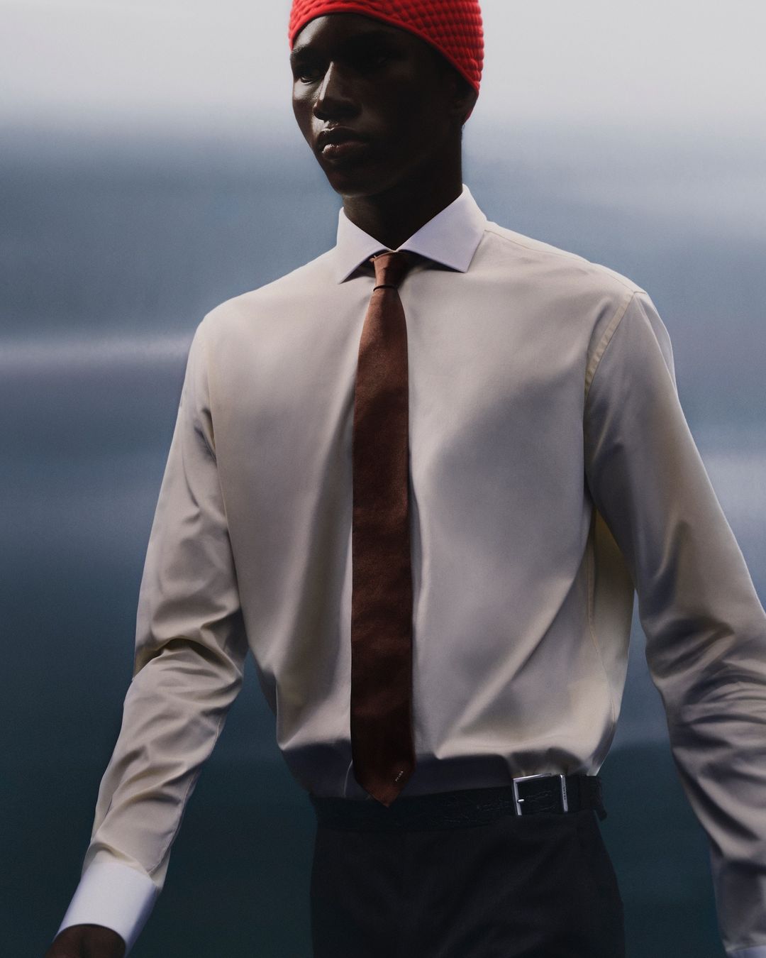 A person wearing a white collared shirt, brown tie, and red knit cap against a blurry background in a fashion style image.