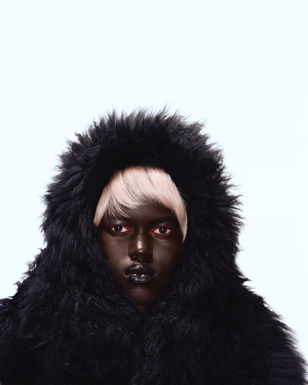 Fashion portrait of a dark-skinned woman wearing a black furry hooded coat with blonde hair on a white background in a photo studio.