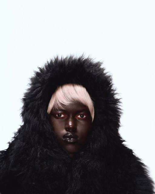Fashion portrait of a dark-skinned woman wearing a black furry hooded coat with blonde hair on a white background in a photo studio.